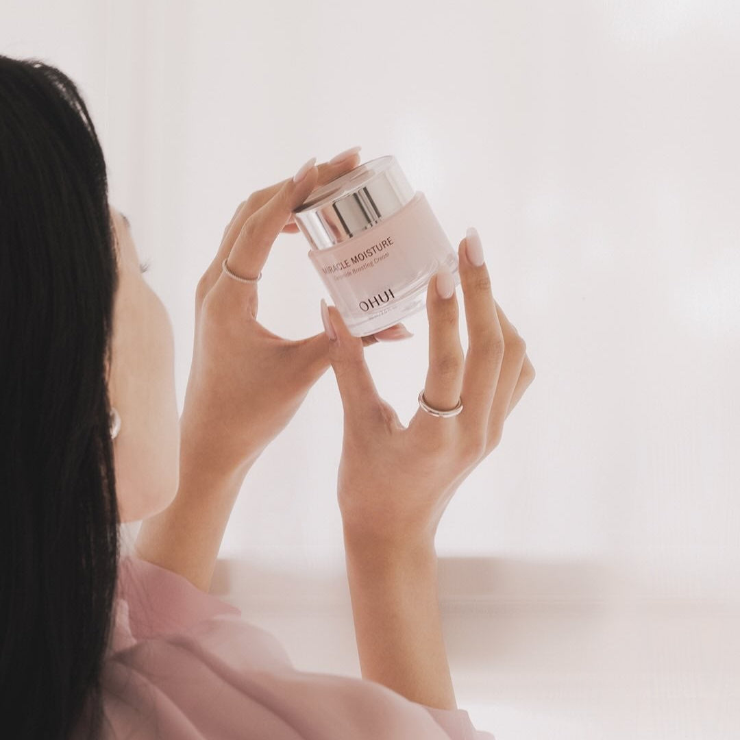 Décris l’image en français : OHUI Miracle Moisture Ceramide Boosting Cream 60ml, marque OHUI, femme tenant le pot en salle de bain.