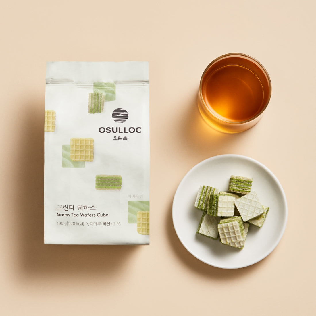 Décris l'image en français : OSULLOC Green Tea Wafers 100g, sachet et gaufres vertes avec tasse de thé sur fond clair.