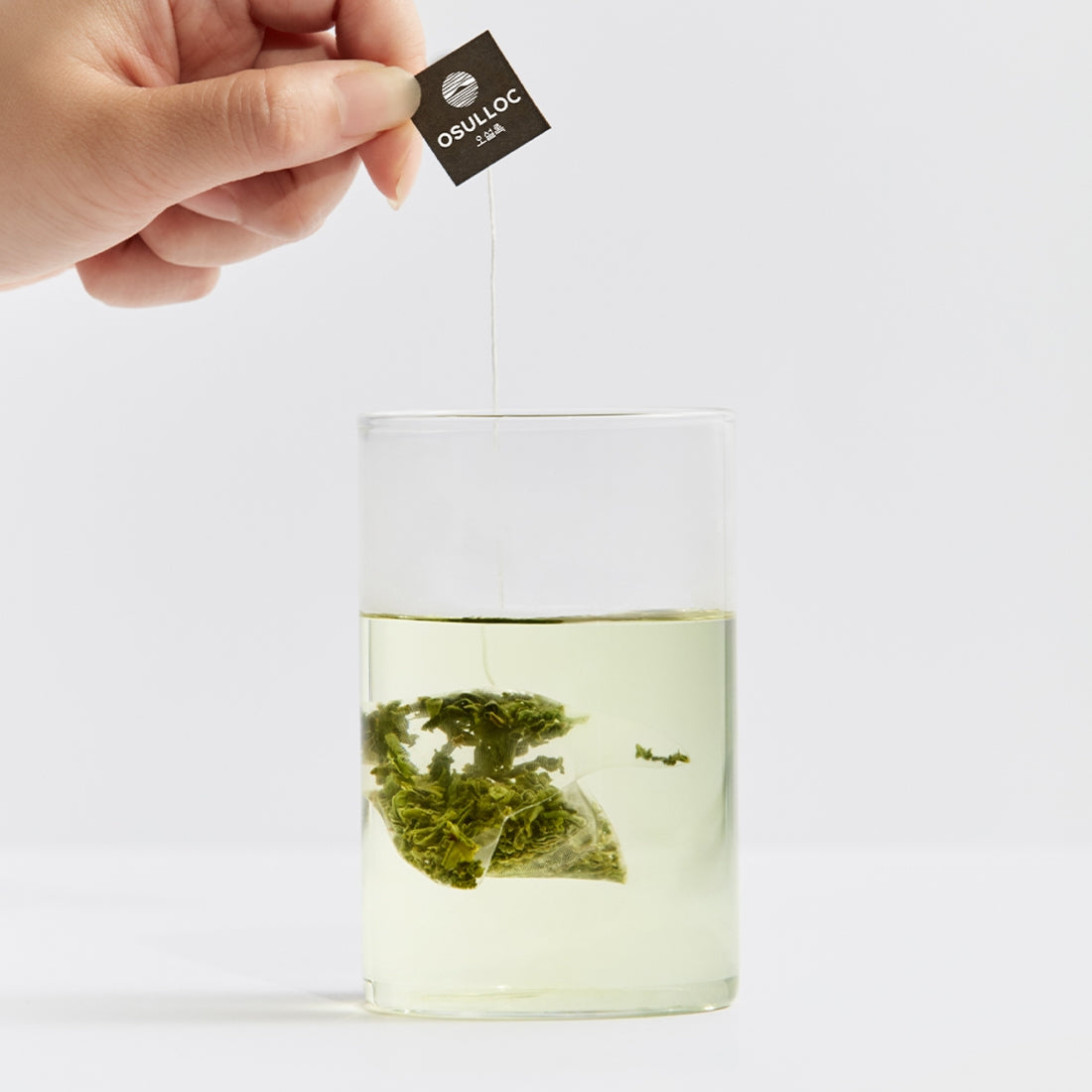 OSULLOC Sejack Green Tea 20 P : sachet vert infusé, marque OSULLOC, main tenant l’étiquette, fond clair, photo produit.