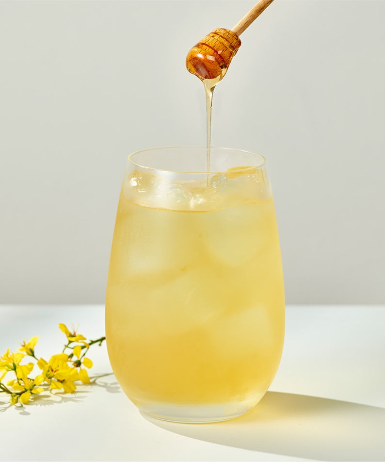 Verre de thé glacé OSULLOC Canola & Apricot Tea 20 P, garni de miel avec des fleurs de canola à côté.