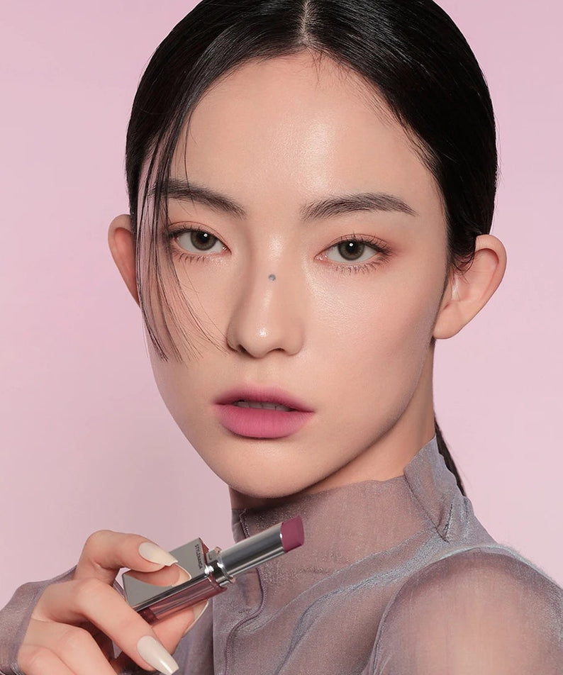 Modèle maquillée appliquant la 3CE Blur Matte Lipstick (3CE) sur fond rose, rendu lèvres mates.