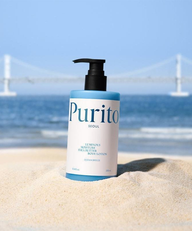 PURITO Luminous Moisture Shea Butter Body Lotion 300ml, flacon sur une plage en bord de mer, produit Purito en contexte visuel.