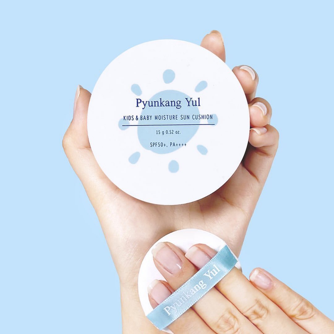 PYUNGKANG YUL Kids & Baby Moisture Sun Cushion SPF50+ PA++++ 15g, boîte blanche tenue par des mains sur fond bleu.