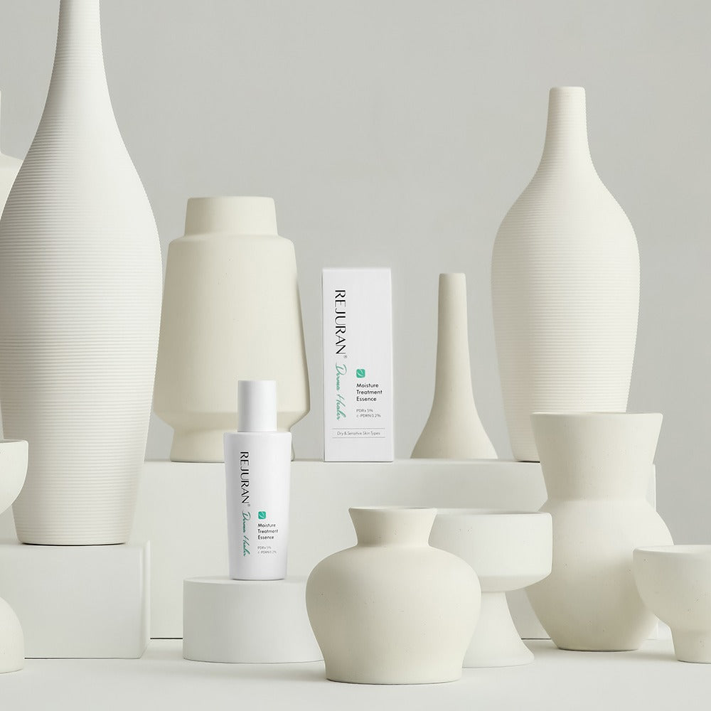 Décris l'image en français : REJURAN Derma Healer Moisture Treatment Essence 70g, flacons blancs sur décor minimal, marque REJURAN.