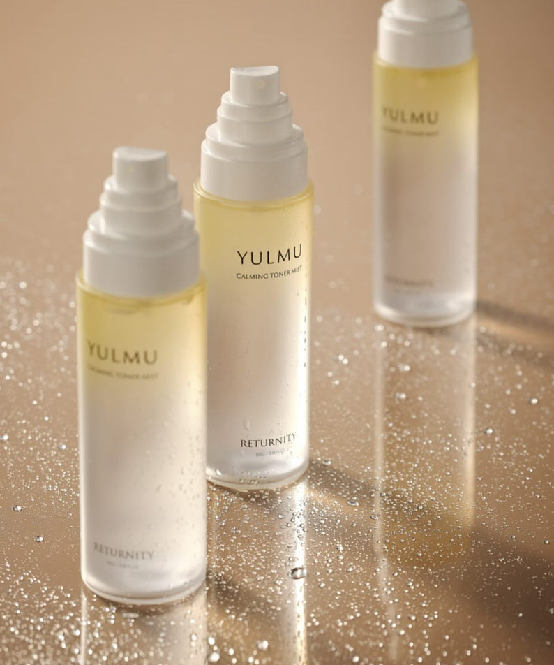 Flacons de RETURNITY Yulmu Calming Toner Mist 100ml brillants sur une surface mouillée, peau apaisée et rafraîchie.