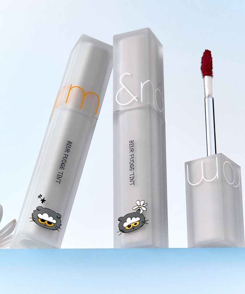 ROMAND x Joanne Friends Blur Fudge Tint, rouge à lèvres liquide élégant sur fond de ciel bleu avec fleurs.