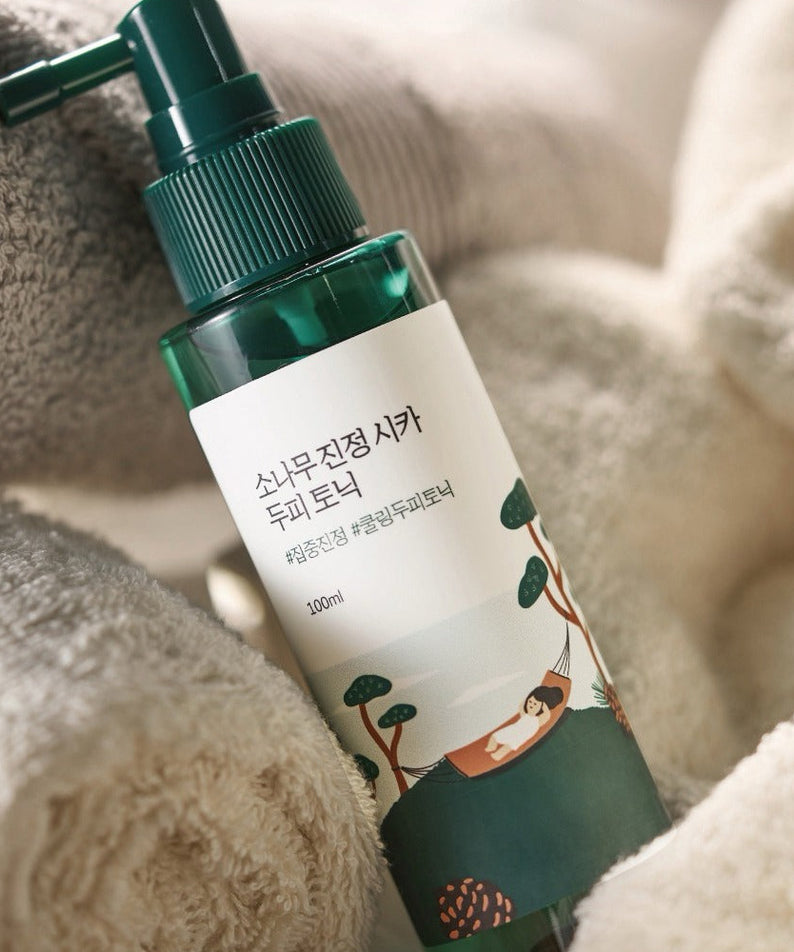 Flacon de ROUND LAB Pine Calming Cica Hair Tonic 100ml au milieu de serviettes douces.