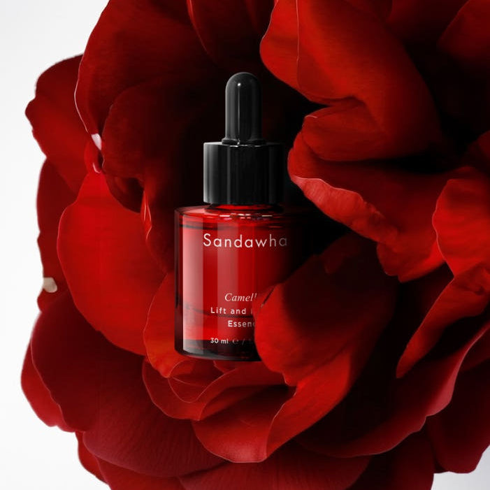 Décris l'image en français : SANDAWHA Camellia Lift & Renew Essence 30ml, flacon noir sur fleurs rouges camélia.