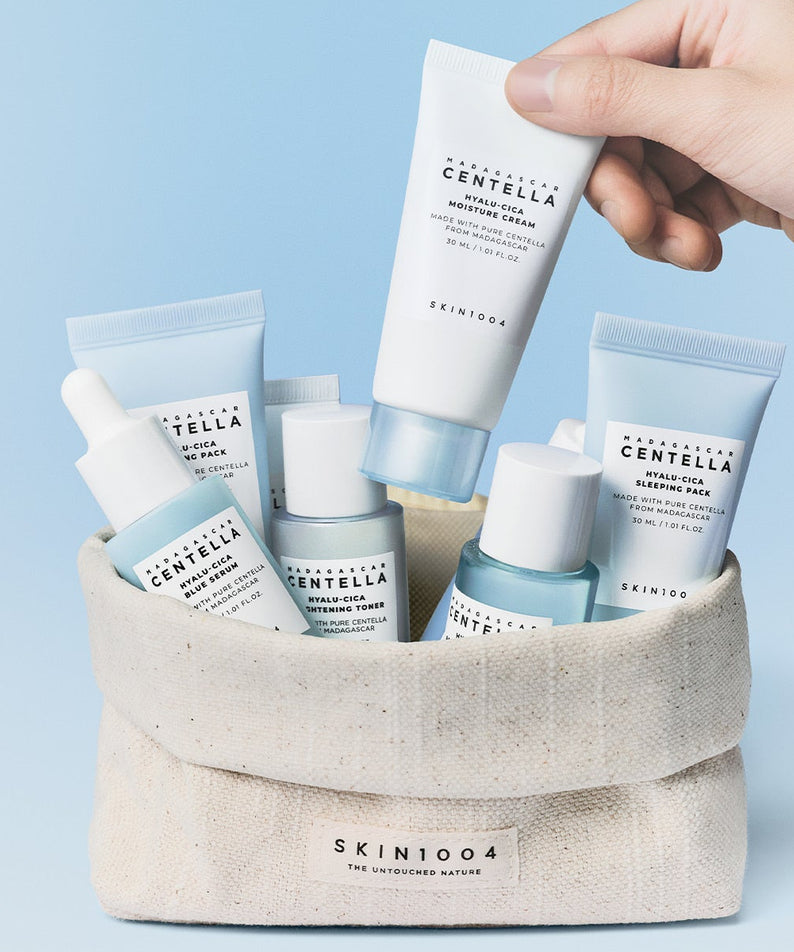 SKIN1004 Madagascar Centella Hyaluronic Acid Travel Kit: produits SKIN1004 en trousse, main tenant un tube, fond bleu.