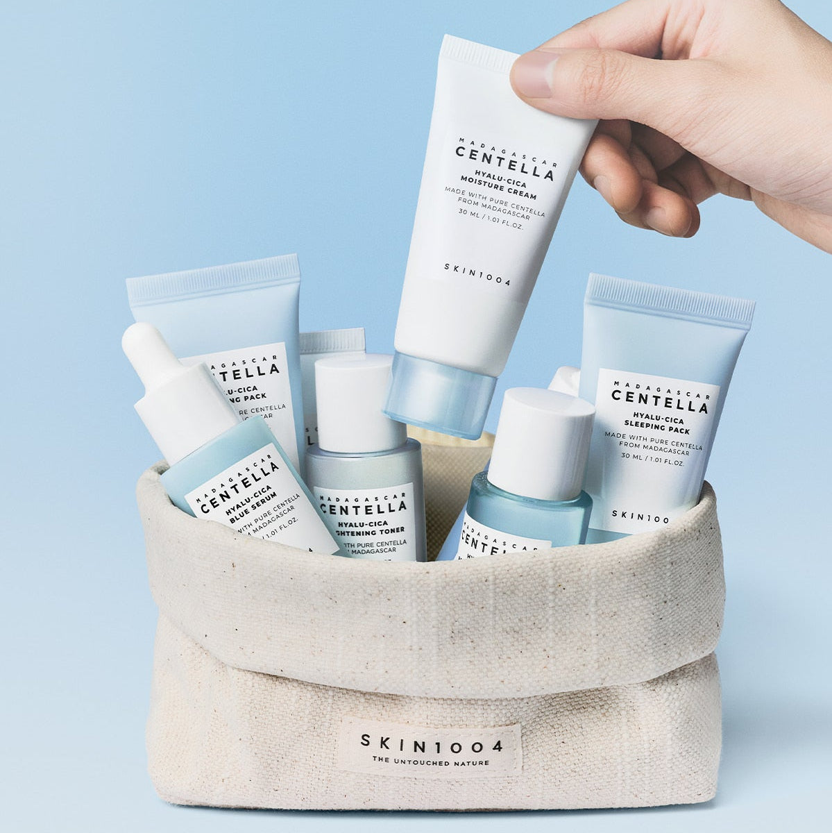 SKIN1004 Madagascar Centella Hyaluronic Acid Travel Kit: produits SKIN1004 en trousse, main tenant un tube, fond bleu.