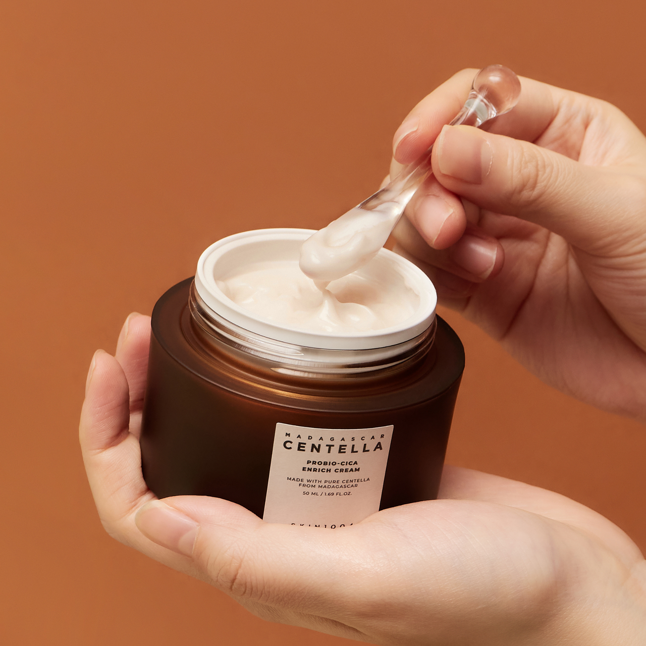 SKIN1004 Madagascar Centella Probio-cica Enrich Cream 50ml : crème sur pot ouverte, main tenant, fond orange, contexte skincare