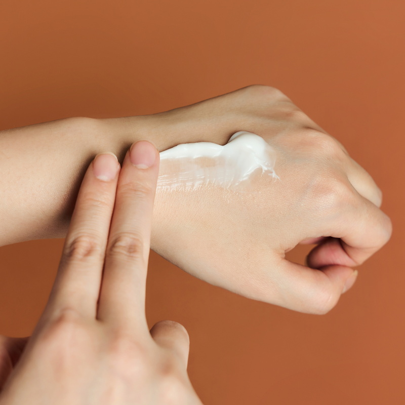 SKIN1004 Madagascar Centella Probio-cica Enrich Cream 50ml : main applique une crème sur peau, contexte orange, flacon non visible