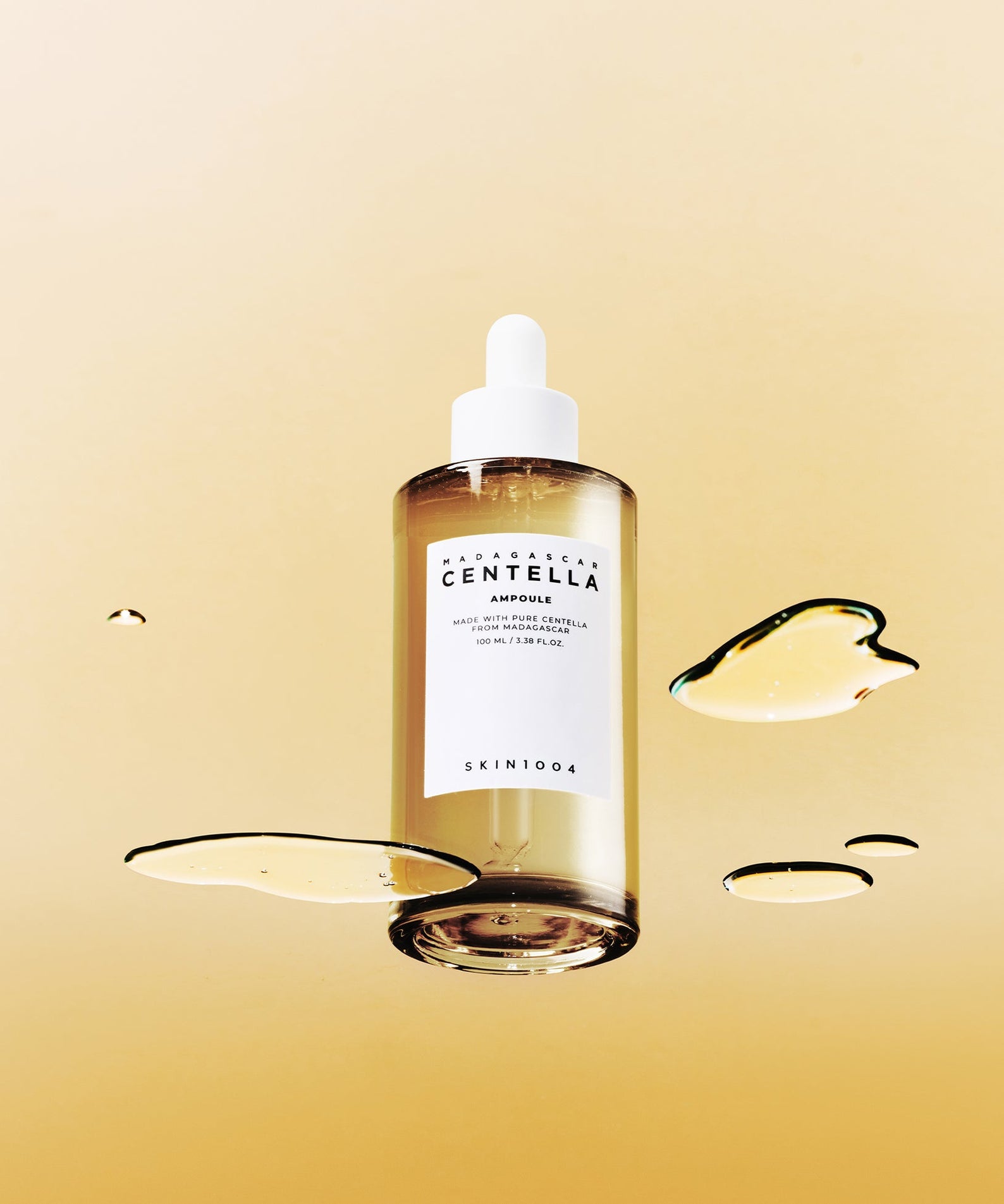 SKIN1004 Madagascar Centella Ampoule 100ml sur fond beige avec gouttes d'ampoule autour, bouteille visible au centre.