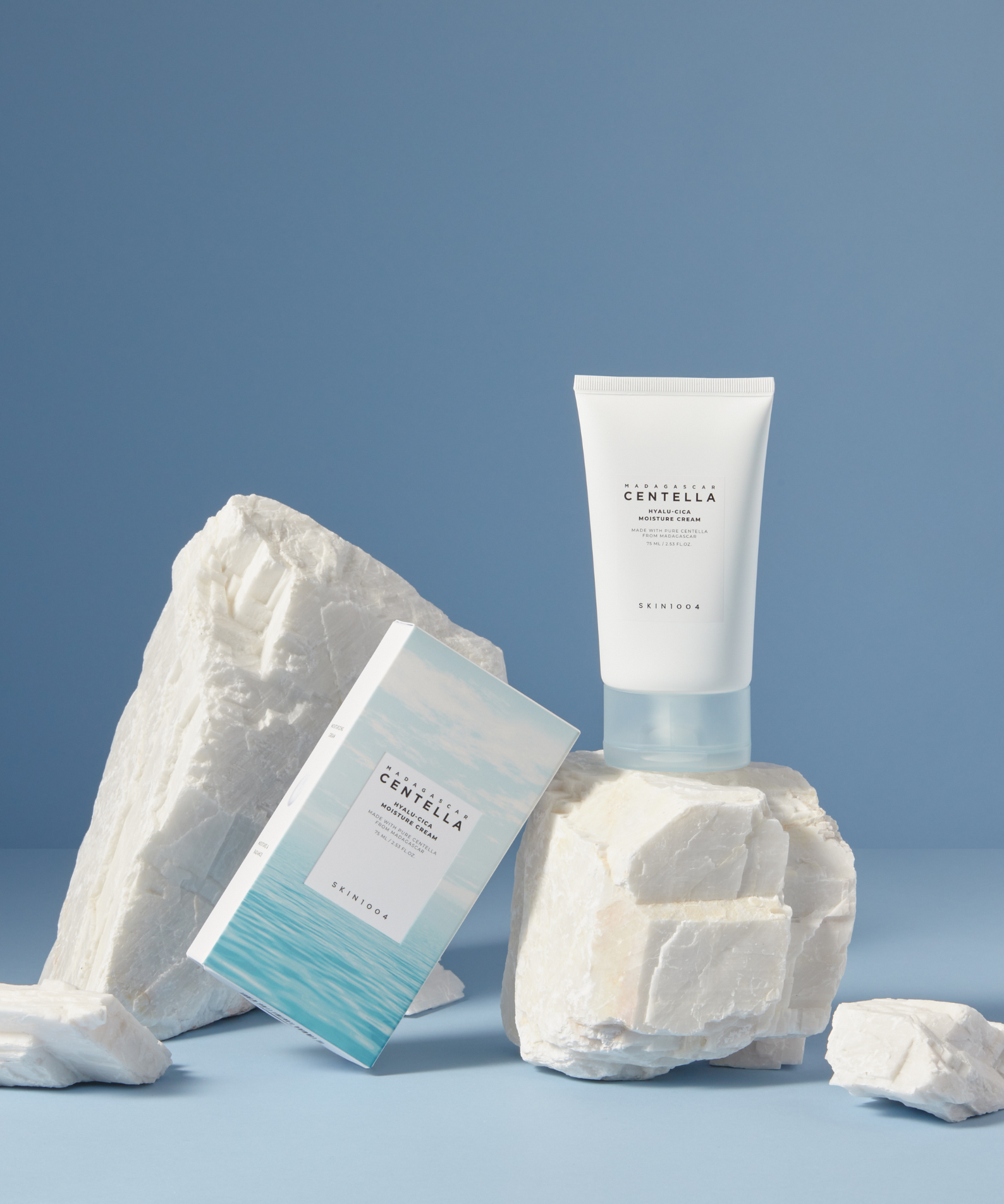 Crème hydratante SKIN1004 Madagascar Centella Hyalu-Cica 75ml avec emballage, posée sur des roches blanches, arrière-plan bleu.