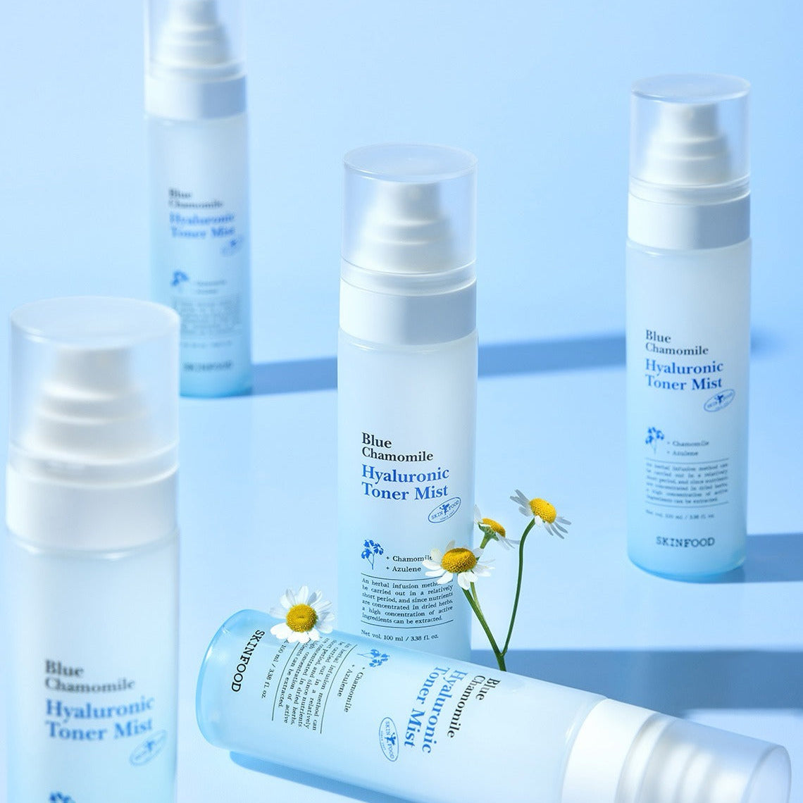 Décris l'image en français: SKINFOOD Blue Chamomile Hyaluronic Toner Mist 100ml, flacons blancs sur fond bleu avec fleurs de camomille.