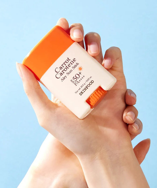 SKINFOOD Carrot Airy Soft Sun Stick 18.5g, marque SKINFOOD, tube crème solaire tenue sur fond bleu clair.
