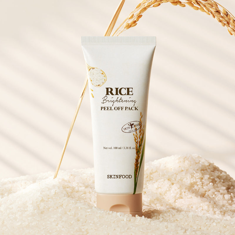 SKINFOOD [NEW] Rice Brightening Peel-Off Pack 100ml, tube crème sur fond beige avec grains de riz et ruban doré