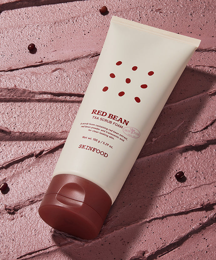 SKINFOOD Red Bean Tranexamic Acid Scrub Foam 150g en tube sur fond rose texturé, marque SKINFOOD, contexte visuel beauté.