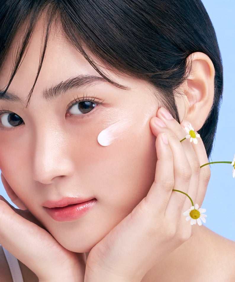 Femme appliquant du SKINFOOD Carrot Airy Soft Sun Stick 18.5g sur la peau, avec des fleurs de camomille, fond bleu.