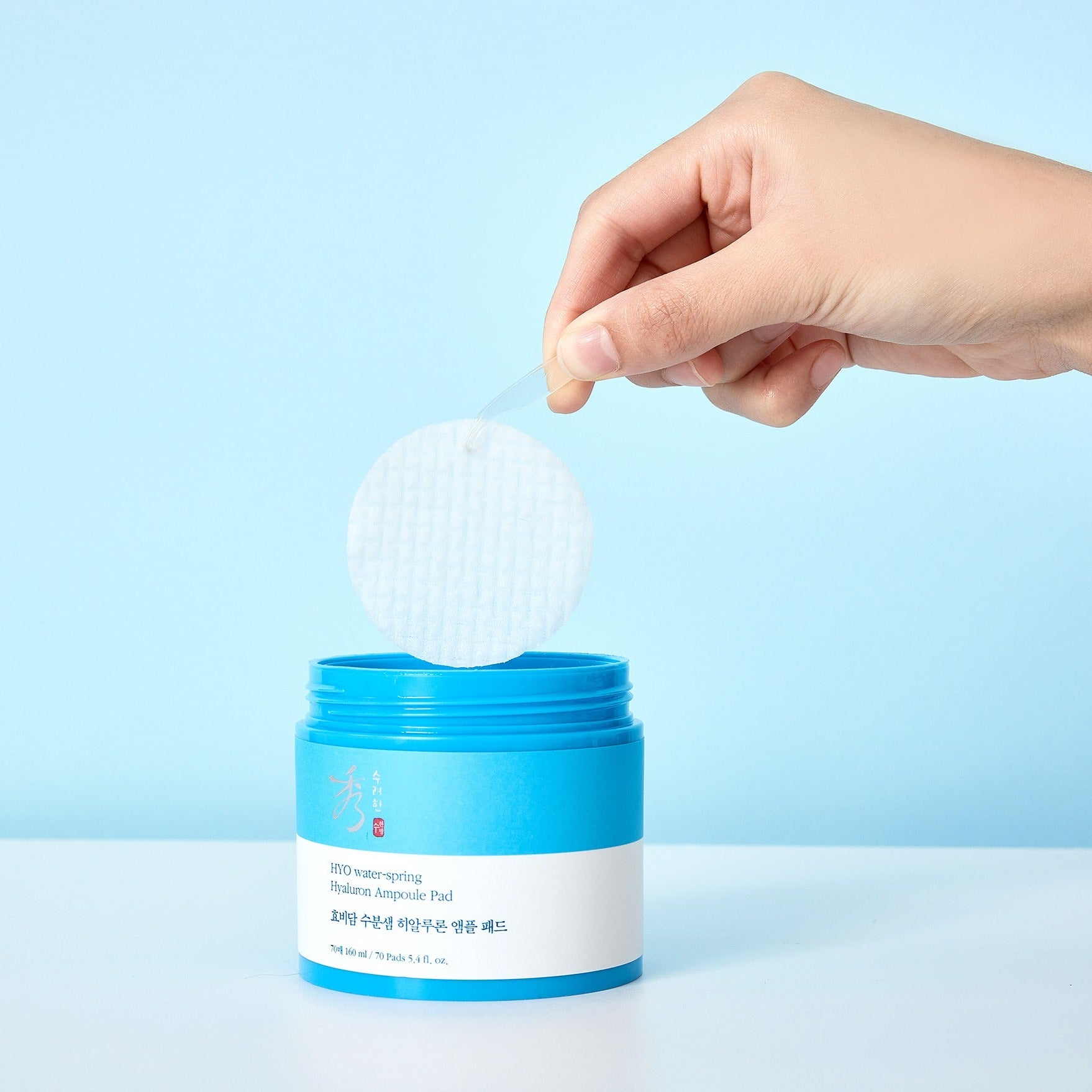 Main tenant un pad hydratant avec SOORYEHAN Moiture Spring Hyaluronic Ampoule Pads 70 Sheets, marque sur le pot bleu.