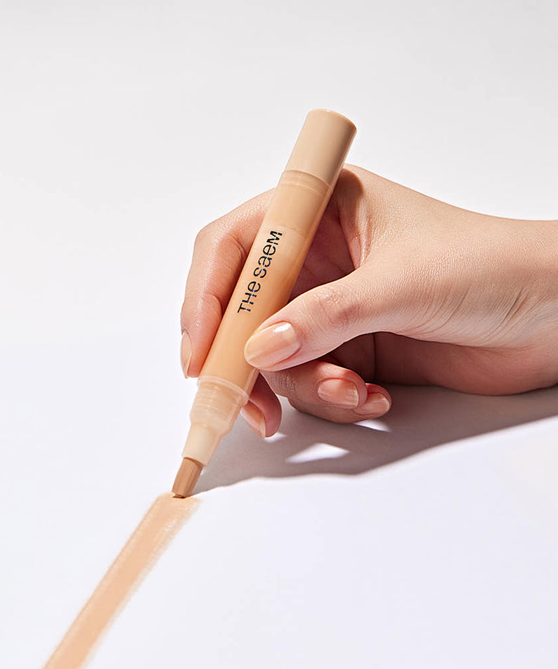 THE SAEM Cover Perfection Pen Concealer : main tient le stylo correcteur maquillage, fond blanc, marque THE SAEM.