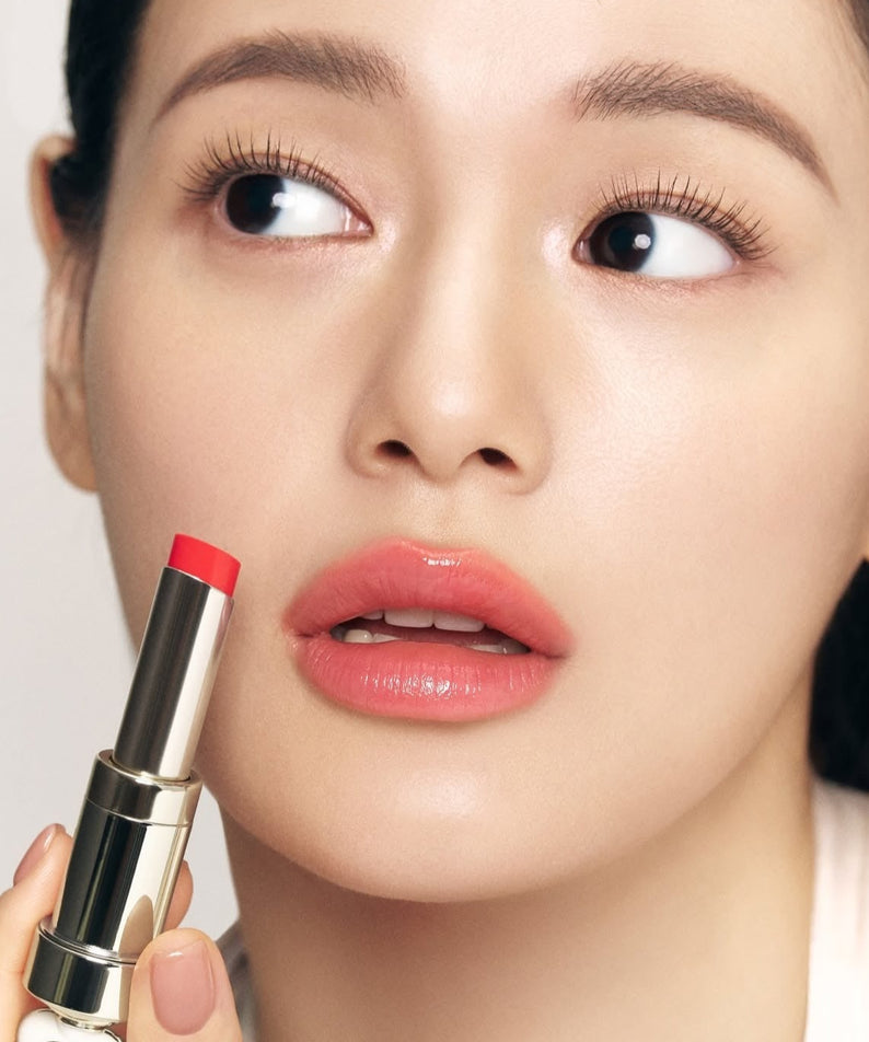 THE WHOO Essentail Lip Glow Balm : femme maquillée tenant un baume à lèvres rouge, gros plan, fond clair.