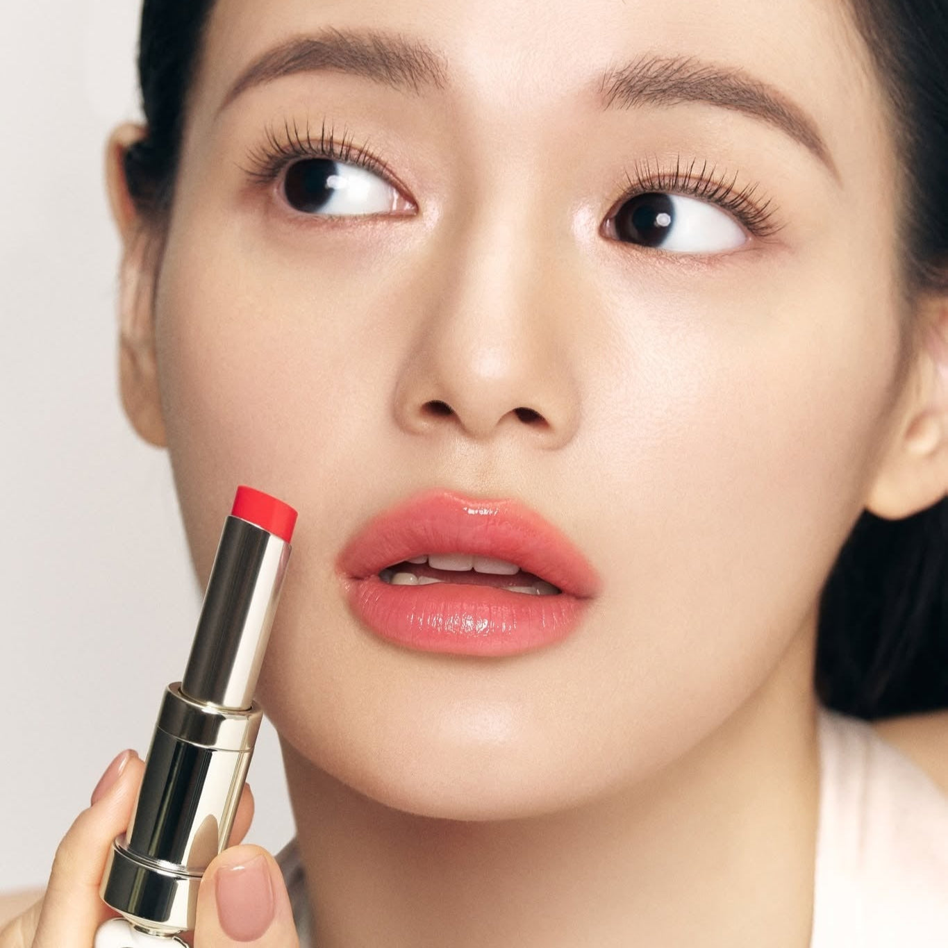 THE WHOO Essentail Lip Glow Balm : femme maquillée tenant un baume à lèvres rouge, gros plan, fond clair.
