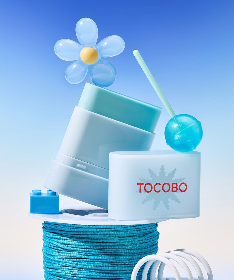 TOCOBO Mini Sun Stick Trio de TOCOBO, en pack sur fond bleu ciel avec accessoires visuels, fleur et soleil stylisés.