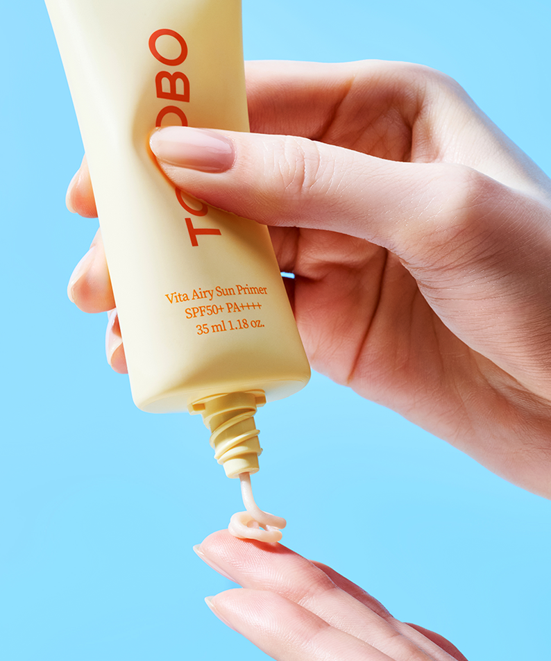 Application du TOCOBO Vita Airy Sun Primer 35ml sur les doigts, produit solaire SPF50 avec une texture légère sur fond bleu.
