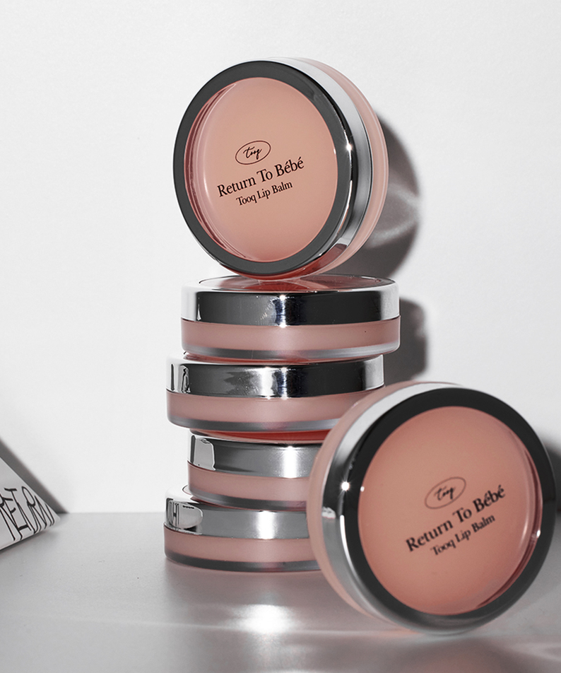Empilement de baumes à lèvres TOOQ Return To Bèbè Lip Balm élégamment disposés, reflets argentés sur fond blanc.