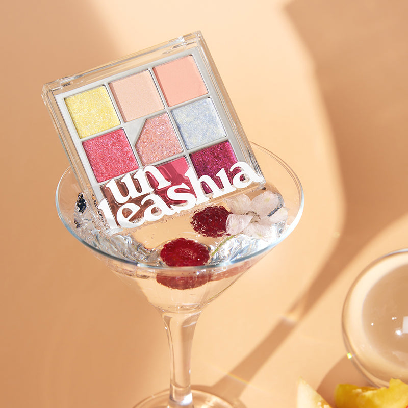 UNLEASHIA Glitterpedia Eye Palette, palette fards pailletés en coffret transparent sur fond beige, gros plan en lumière chaude.
