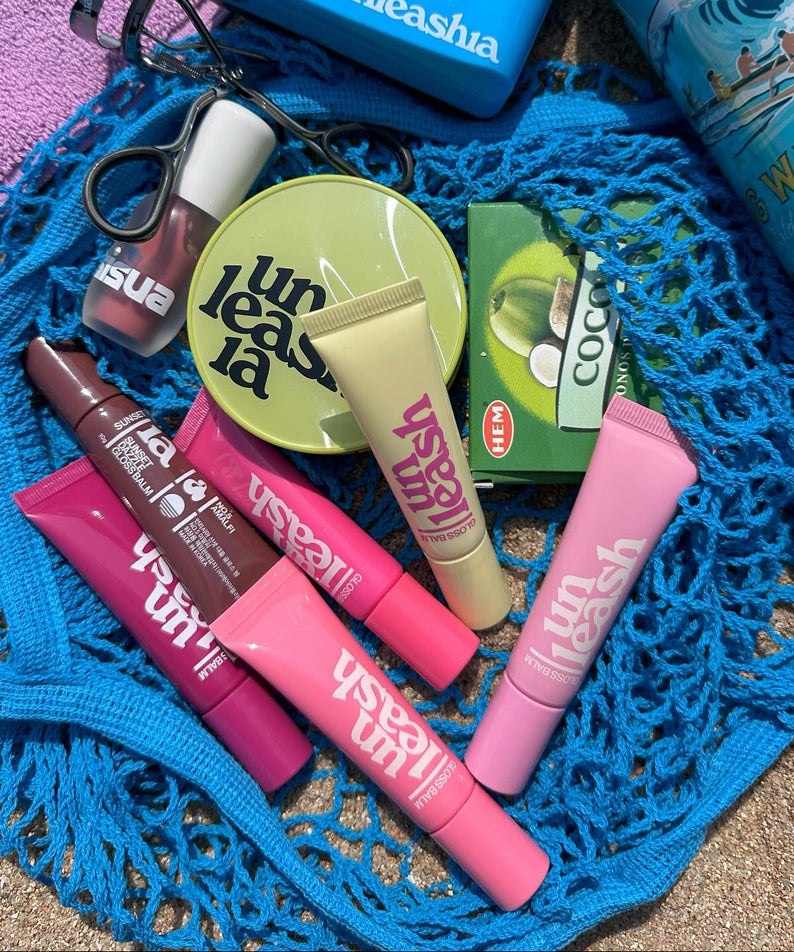 Produits cosmétiques colorés UNLEASHIA Sunset Dazzle Gloss Balm sur une nappe bleue, ambiance estivale.