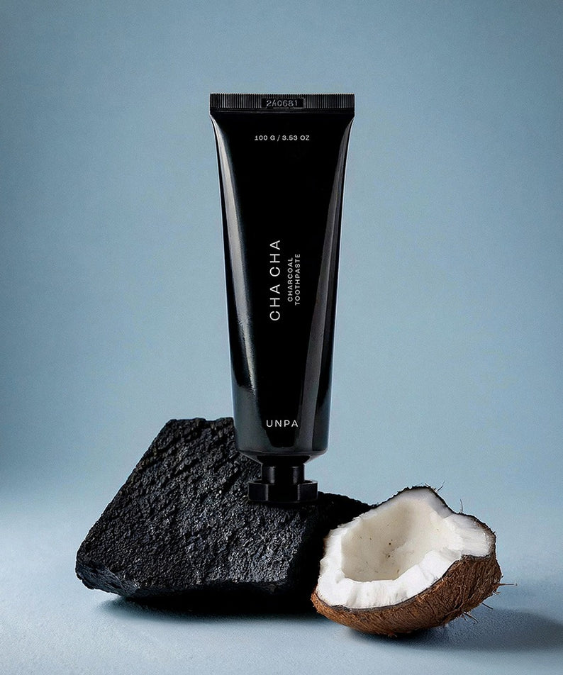 UNPA Cha Cha Charcoal Original Toothpaste 100g de UNPA : tube noir sur charbon et noix de coco, fond bleu.