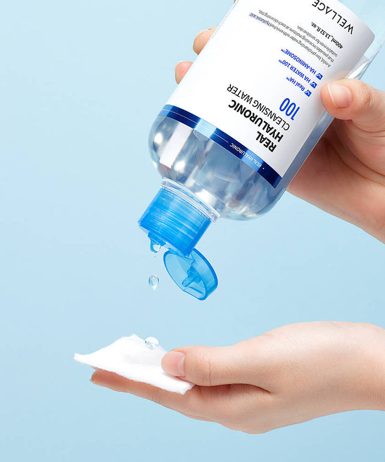 WELLAGE  Real Hyaluronic Cleansing Water 400ml, main tenant le flacon avec goutte sur peau, fond bleu ciel