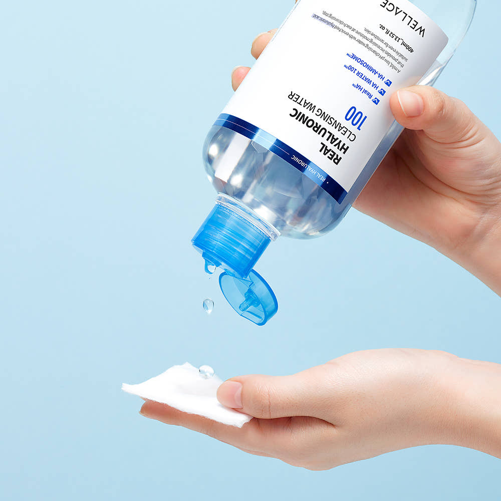 WELLAGE  Real Hyaluronic Cleansing Water 400ml, main tenant le flacon avec goutte sur peau, fond bleu ciel