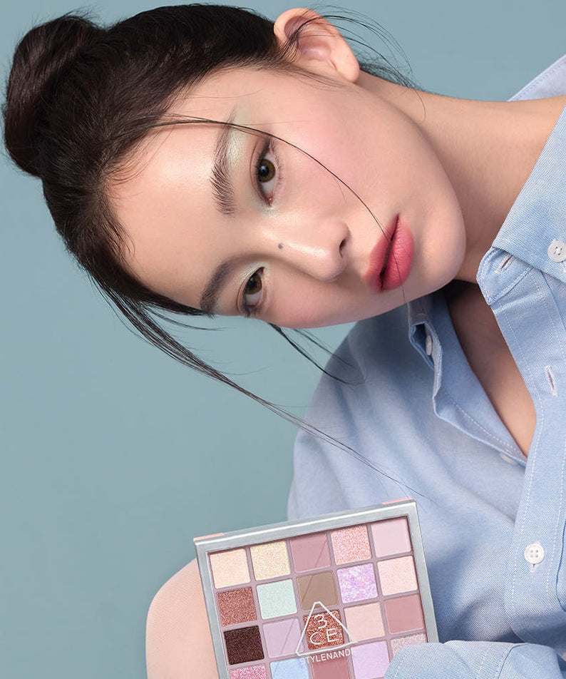 Femme posant avec la 3CE XL Eyeshadow Palette, palette de fards à paupières, sur fond bleu, mettant en valeur ses teintes variées.