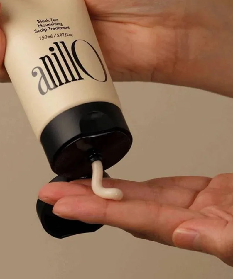 Décris l'image en français : ANILLO Black Tea Nourishing Scalp Treatment 150ml, tube tenu par une main, crème appliquée sur cuir chevelu.