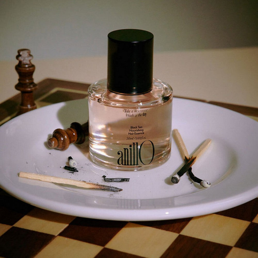 ANILLO Black Tea Nourishing Hair Essence 50ml : flacon noir et ambre sur une assiette, décor salon cosmétique, ambiance naturelle.
