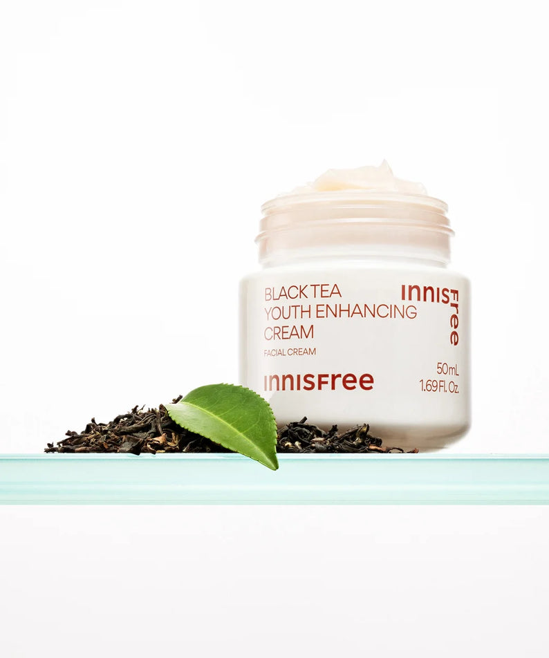 Décris l'image en français : INNISFREE Black Tea Youth Enhancing Cream 50ml, pot de crème sur fond blanc avec feuilles de thé noir.