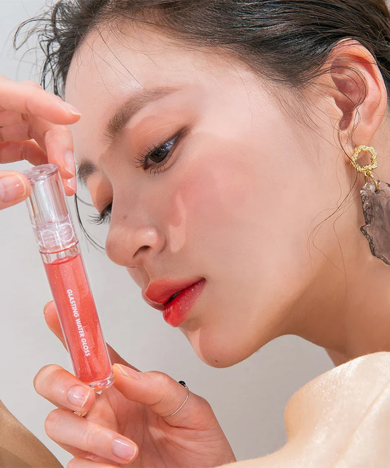 ROMAND Glasting Water Gloss, flacon rosé appliqué sur le maquillage, gros plan visage en studio, main à côté.